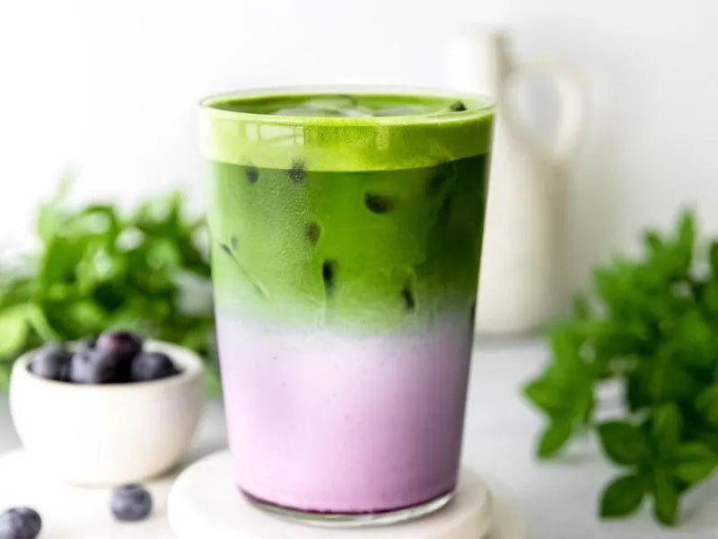 Layered blueberry matcha latte in a clear glass with blueberry purée, milk, and ceremonial matcha on a neutral beige background.